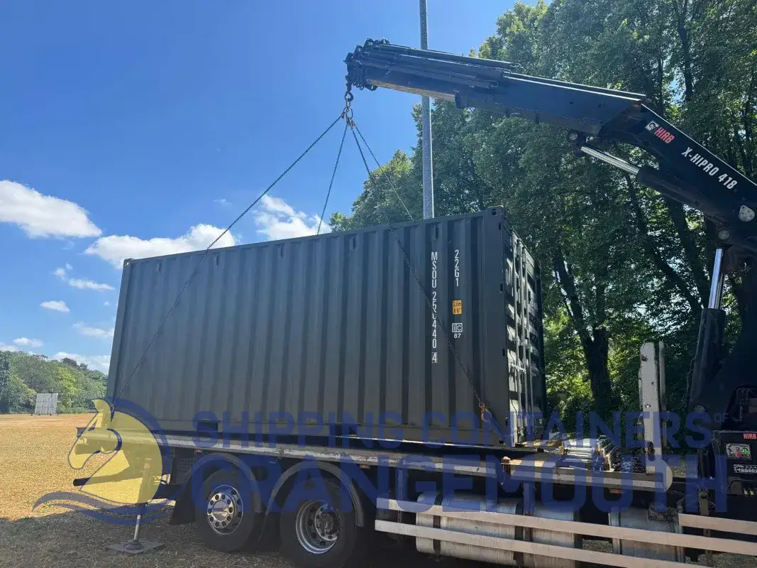 A crane lifts a shipping container above a parked truck under a blue sky with scattered clouds.