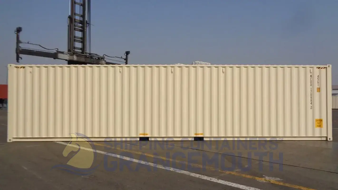 A beige shipping container stands on a dock, with cranes and other containers visible in the background.